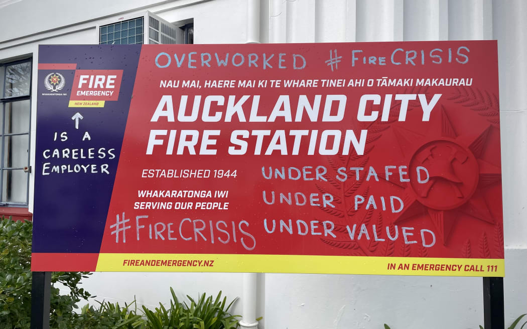 A sign outside the Auckland city fire station. Over the sign, protest graffiti reads 'OVERWORKED - #FIRECRISIS - UNDER STAFFED - UNDER PAID - UNDER VALUED'. Next to the logo for Fire and Emergency New Zealand, there is an arrow saying 'IS A CARELESS EMPLOYER'.