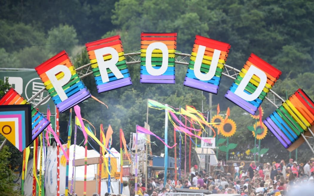 Festivalgoers walk under a rainbow-coloured "Proud" sign at the Glastonbury festival near the village of Pilton in Somerset, southwest England, on June 23, 2022. - More than 200,000 music fans and megastars Paul McCartney, Billie Eilish and Kendrick Lamar descend on the English countryside this week as Glastonbury Festival returns after a three-year hiatus. The coronavirus pandemic forced organisers to cancel the last two years' events, and those going this year face an arduous journey battling three days of major rail strikes across the country. (Photo by ANDY BUCHANAN / AFP)