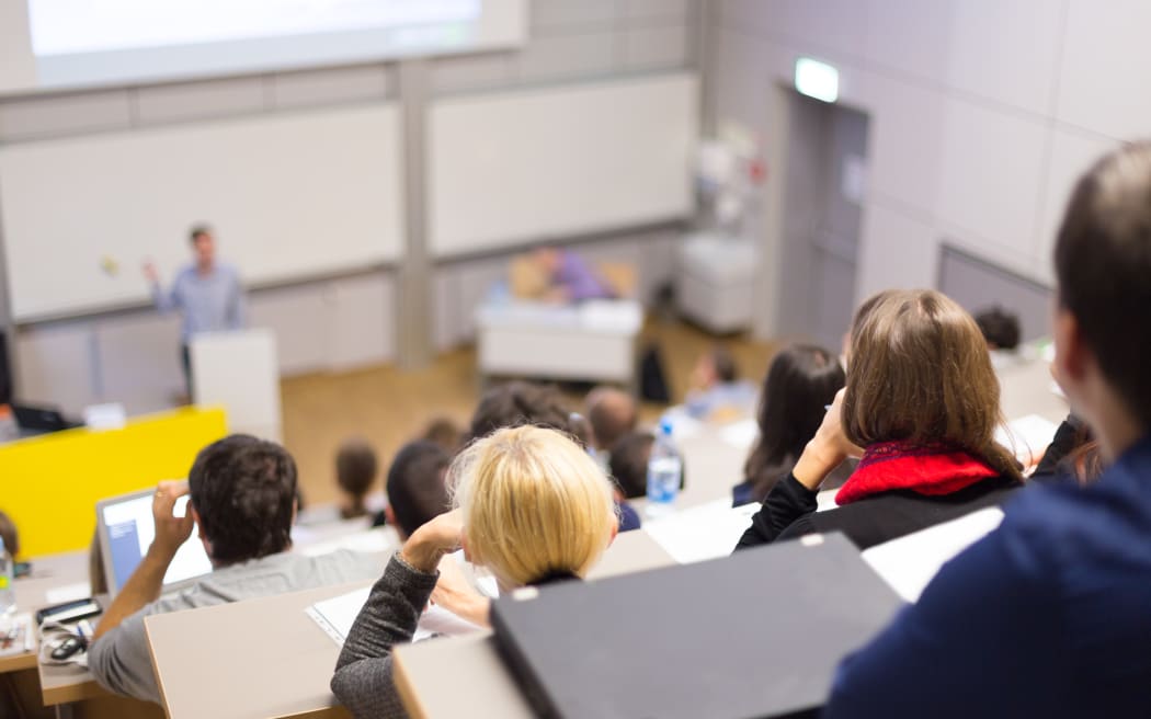 Professor giving presentation in lecture hall at university. Participants listening to lecture and making notes.