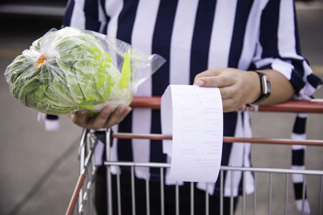 Woman shopping with trolley, holding receipt of grocery items, with food in handing, checking prices.