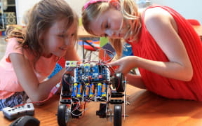 Two primary aged girls explore a wheeled battery powered robot on a table