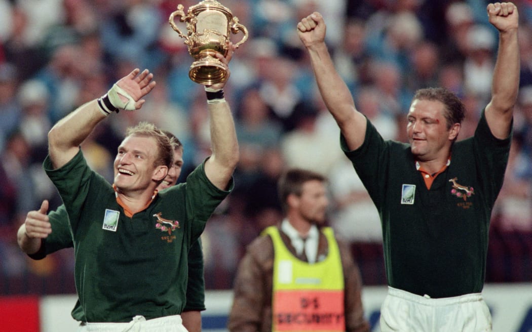 South African captain François Pienaar brandishing the William Webb Ellis trophy and saluting the crowd with teammate Hannes Strydom after beating New Zealand to win the 1995 Rugby World Cup final.