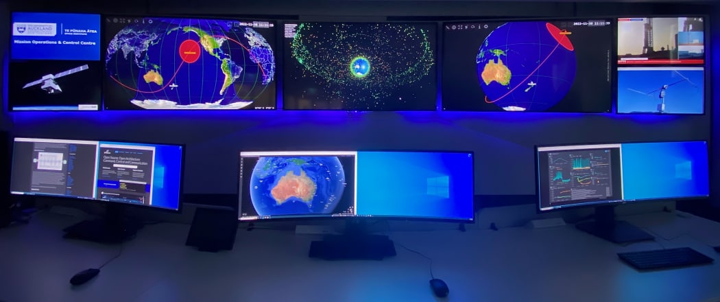 A wide angle shot of the control centre showing two rows of monitors, 11 in total, with large ones on top and smaller below. They are displaying maps of the earth, satellite orbits, satellites, rocket launches, and some blank blue screens.