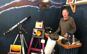 Paul Bishop stands behind a small desk. To the left of Paul is a collection of star-gazing equipment, including telescopes, as well as framed pictures of stars.