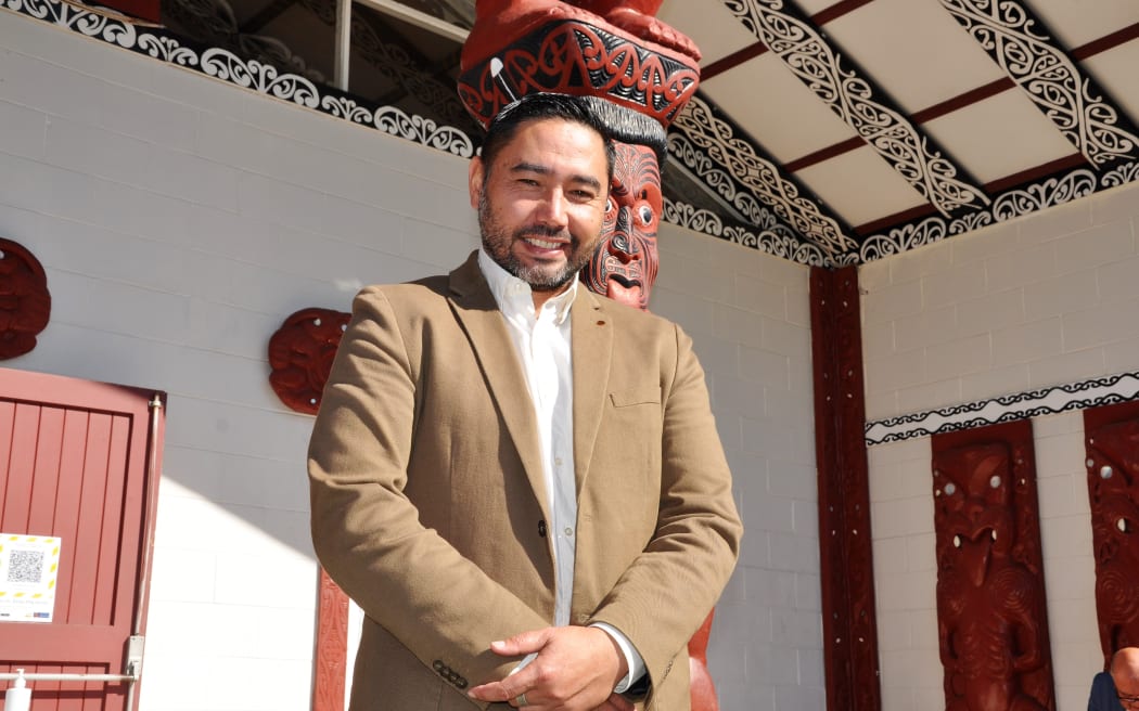 Te Matapihi general manager Wayne Knox stands in front of a wharenui wearing a white collared button down shirt and beige blazer.