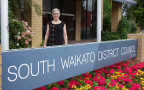 Female mayor stands behind a council sign with bright municpal flowers planted in front