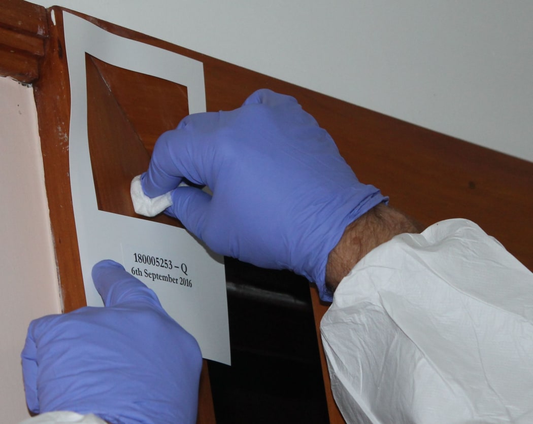 A close up photo of two hands with gloves on, swabbing within a 100 square centimetre stencil stuck to a wall. By swabbing within that area, testing companies get a sample that corresponds to Ministry of Health guidelines for meth.