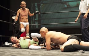 Shintaro Yano ( "Sambo" Shintaro) watches on as tag-team partner L'Amant delivers a kick to able-bodied "Antithesis" Kitajima's face.