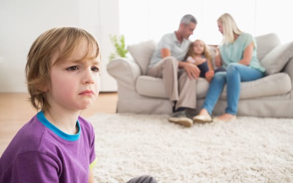 Photo of an upset boy sitting on floor while parents enjoying with sister on sofa at home