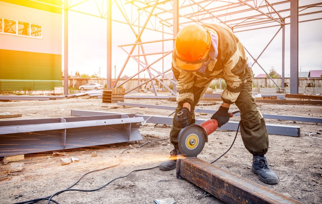 A strong  man welder in brown uniform, a construction helmet and welders leathers, grinder metal an angle grinder  at the construction site, orange sparks fly to the sides