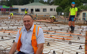 Man sits at front of new concrete foundation with reinforcing with young  apprentice in hihg vis and hard hat standing  behind.