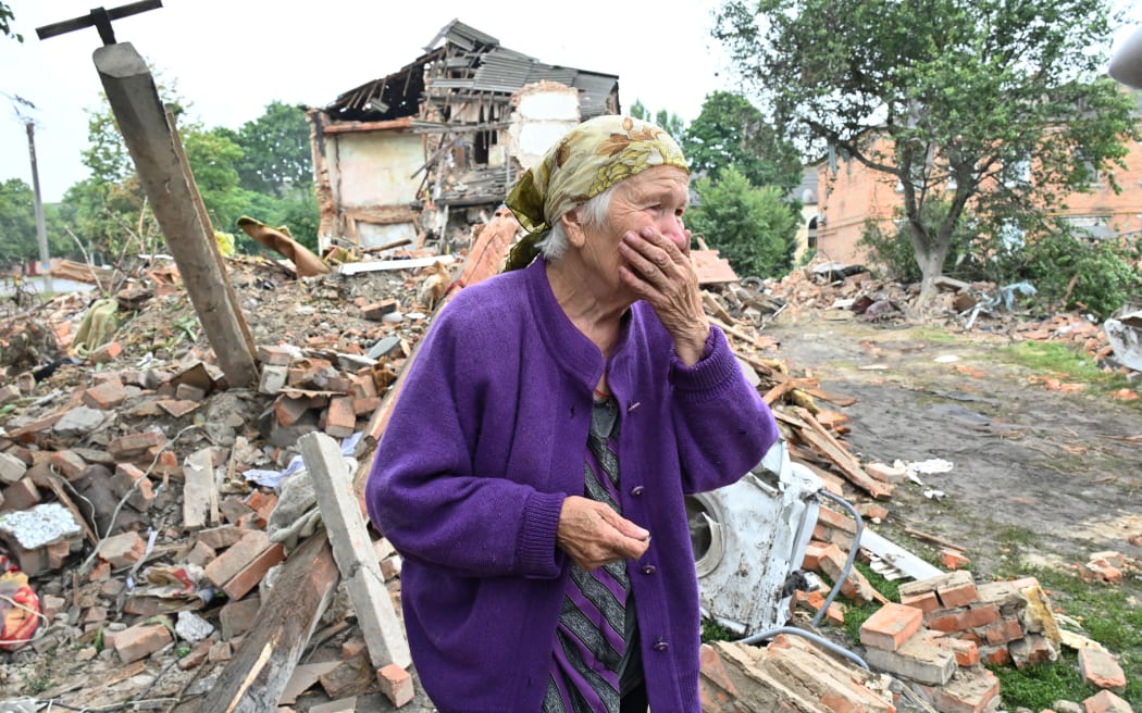 A local resident, Raisa Kuval, 82, reacts next to a damaged building partially destroyed after a shelling in the city of Chuguiv, east of Kharkiv, on July 16, 2022. - In the northeast region around Ukraine's second city of Kharkiv, governor Oleg Synegubov said an overnight Russian missile attack killed three people in the town of Chuguiv. (Photo by SERGEY BOBOK / AFP)
