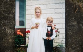 An old photograph. Two blonde children, an older girl and a younger boy, smile at the camera. They are dressed formally, the girl in a white dress holding a bouquet and the boy in a suit with a tartan vest and black bowtie.
