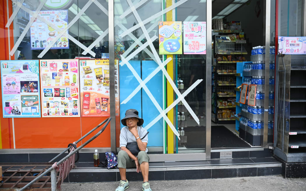 2023 年 9 月 1 日，香港大屿山，一名妇女坐在一家便利店外，窗户贴着胶带进行保护，此时距离超强台风“莎拉”预计抵达的时间还有几个小时。 9 月 1 日，随着超强台风“萨乌拉”席卷中国南部海岸，香港停飞航班、关闭股市并关闭学校。 （彼得·帕克斯/法新社拍摄）