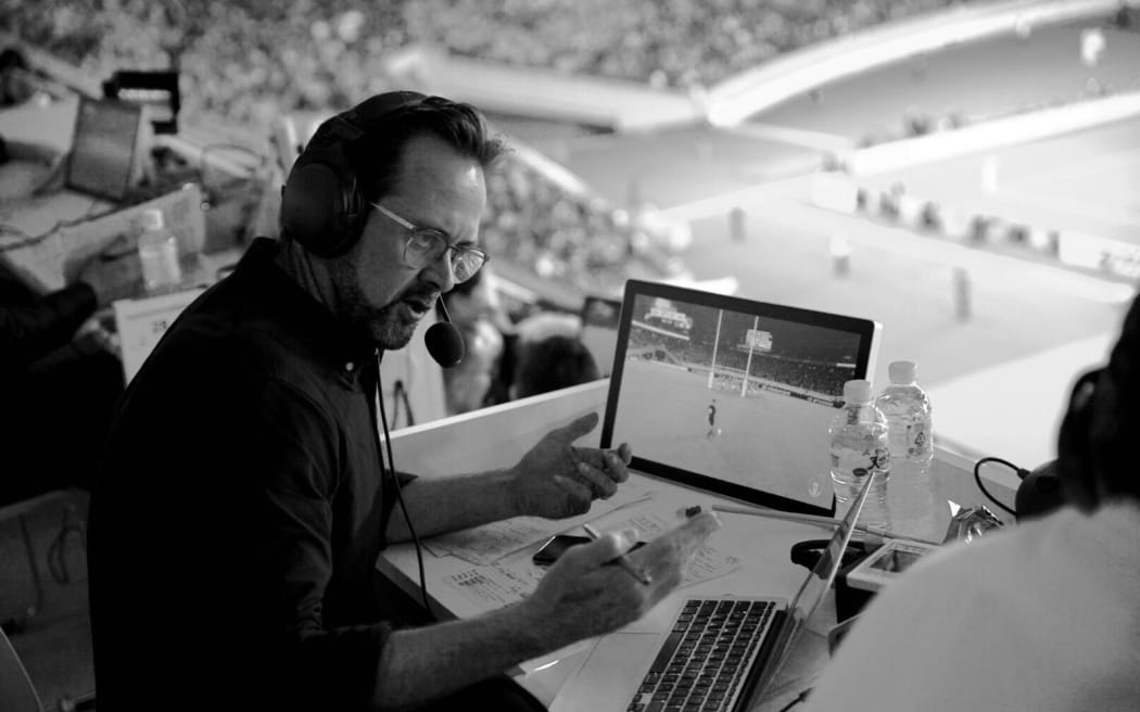 Scotty Stevenson commentates from a desk at the top of a large stadium. He has a screen and a laptop in front of him. He is talking animatedly.
