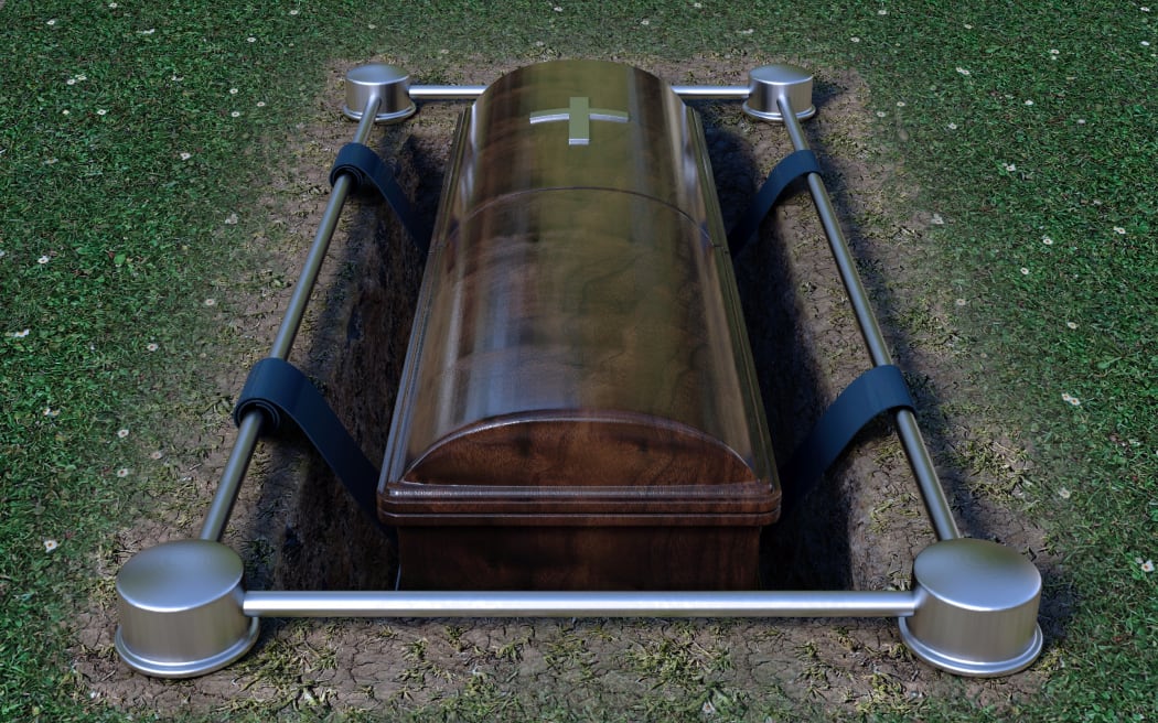 A modern wooden coffin at a funeral being lowered into a grave with a lowering mechanism a dirt and grass background.