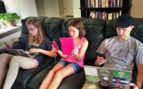 A photo of three children on a couch, all using multiple devices