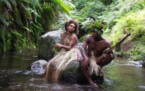 A man and a woman in tradional Ni-Vanuatu clothing sit on a rock in a river