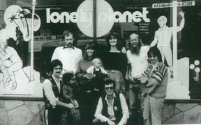 A black and white photograph. A group of people are gathered outside a glass store front. The window says "LONELY PLANET". The people are wearing seventies clothes - stripy woolen knits, vests, pointy collars, and overalls.