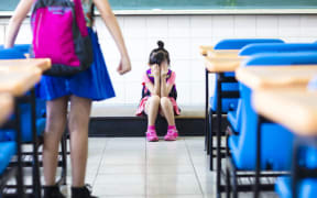 A photo of a girl sitting on her own on the floor of a classroom while all around her other pupils appear to be ignoring her.
