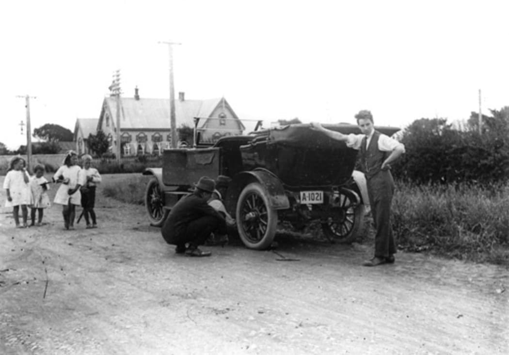 A man changing the flat tyre on his car while a group of small children look on. C. 1920 - 1930.