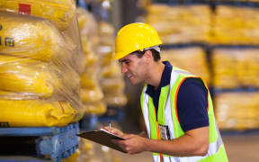A warehouse worker wearing a high-vis vest holds a clipboard (file).