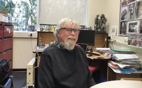 Warren Tate sitting at a desk in his office with photos on the wall and files on the top of shelves and tables behind him.