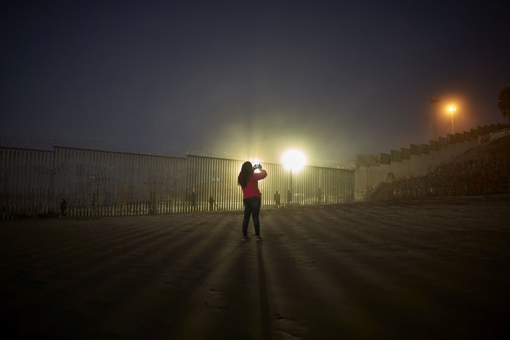 A woman records with her phone, as floodlights from the United States light up the border wall, topped with razor wire, along the beach in Tijuana, Mexico.