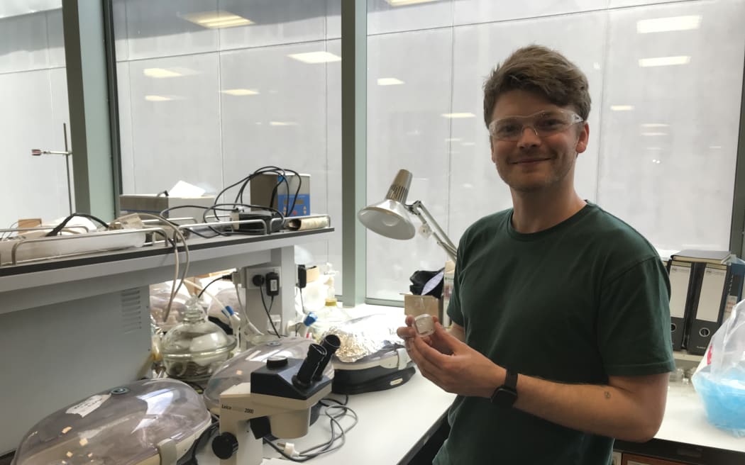 Joe is standing at a lab bench in front of a microscope. He's smiling and holding a sample of specialised material.
