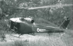 An RNZAF Iroquois hovering just above the ground as it waits to pick up a patrol in the Tilomar area. The door gunner, LAC Daryl Fell, is checking the tail rotor clearance; March 2000.