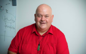 A portrait photo of wellington scientist professor Rod Badcock. We is wearing a red shirt and a pounamu toki necklace.