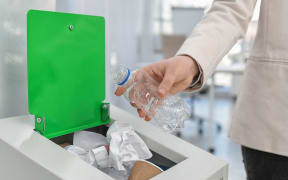 Woman putting used plastic bottle into trash bin in modern office, closeup. Waste recycling