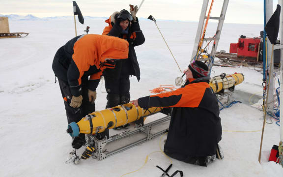 Three scientists in Antarctica, wearing black and orange jackets. They surround a yellow piece of equipment.