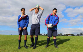 Three teenagers stand with rugby  balls on a field.