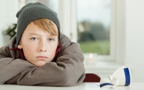 Close Up of Young Teenage Boy Wearing Hoodie and Winter Cap Looking Remorseful While Leaning on Arms in Kitchen with Broken Mug on Table
