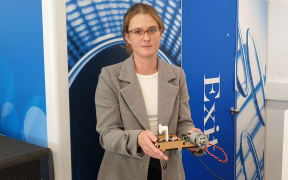 A woman in a blazer and glasses stands in front of a blue sign holding a mechanical device.
