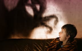 A photo of a boy in the dark with a shadow behind him in the shape of a monster with its arms extended.