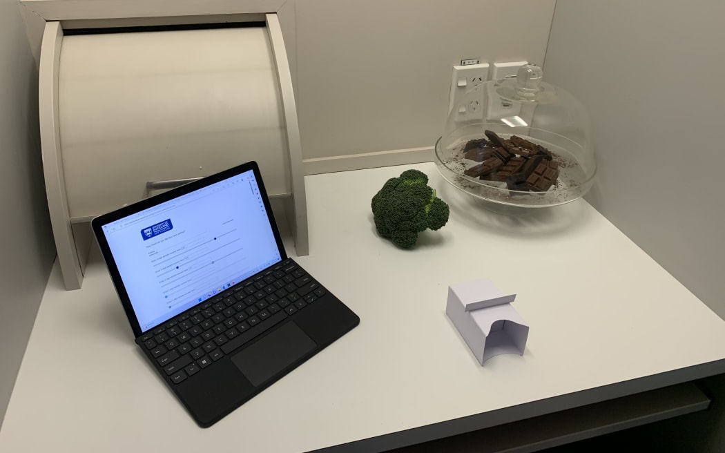 The photo shows a small white cubicle with a serving hatch in the wall behind it. In front of the hatch sits an open laptop showing a programme for rating the test food, a head of raw broccoli, several cubes of dark chocolate under a glass cloche, and a small sealed box used for delivering the mystery test food to the subject while preventing them from either seeing or smelling it.