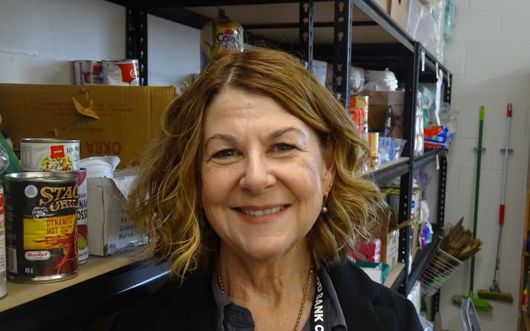Sophie Gray smiles at the camera. She is standing in front of fully stocked shelves of food at the Good Works Trust food bank.