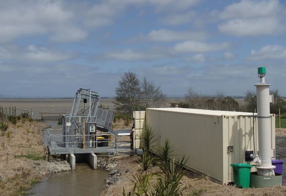A large cage sitting in a small muddy stream, with a shipping container next to it.