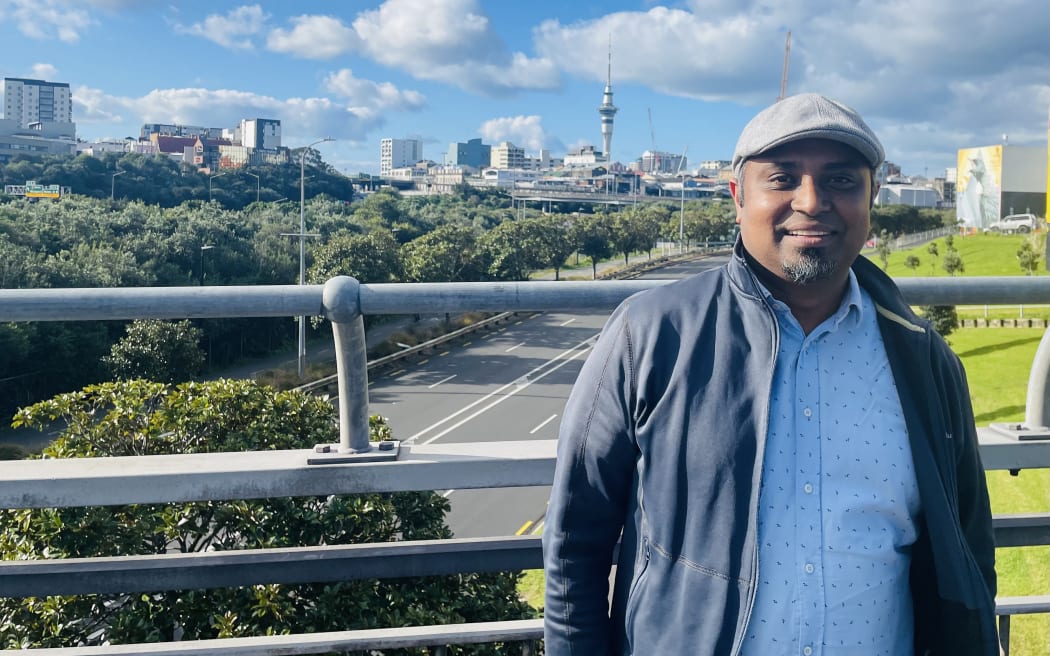 Matthew Prasad smiles at the camera. He is standing on the footpath of the Newton bridge. The Sky Tower is in the background. It is a sunny day with blue skies.