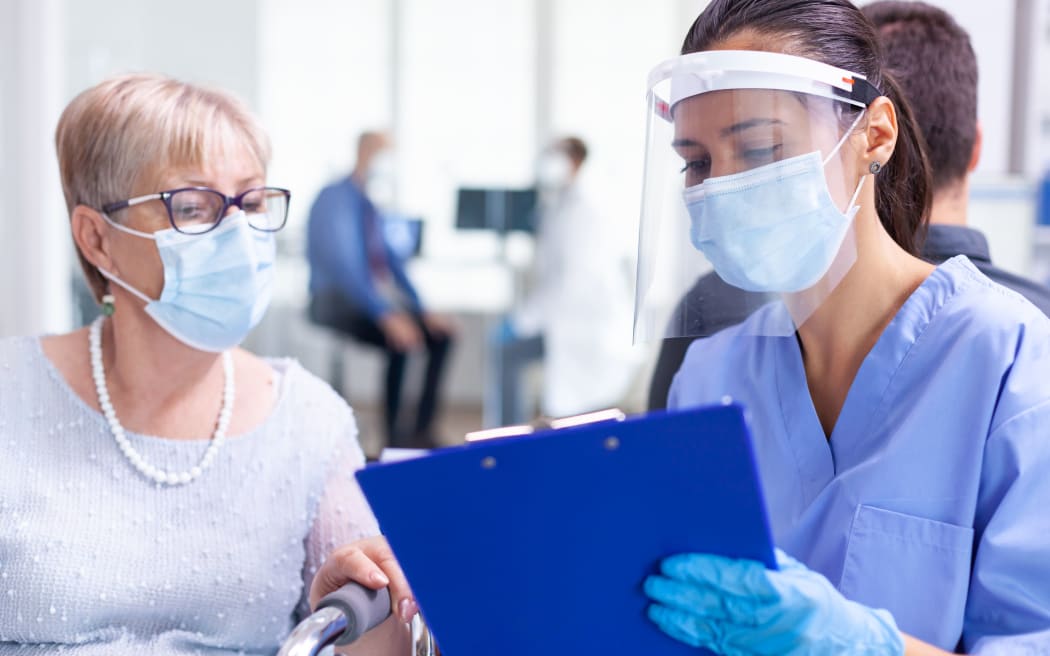 Medical staff wearing visor against coronavirus in hospital hallway talking disabled senior woman wearing face mask. Patient and medical staff in waiting area. Doctor in examination room.