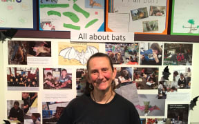 Catriona Gower stands in front of one of the school displays at the Catlins Bats on the Map exhibition. There are photographs and drawings of bats and information about bats on the wall behind her.