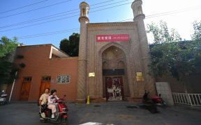 This picture taken on July 13, 2023, shows two people riding a scooter pass a locked mosque bearing the slogan "Love the Communist Party, love the country" in Kashgar, northwestern China's Xinjiang region. Chinese travellers throng the bazaars of old Kashgar, munching mutton kebabs and soaking up heavily commodified Uyghur culture -- part of a government push to remould troubled Xinjiang into a tourism paradise.