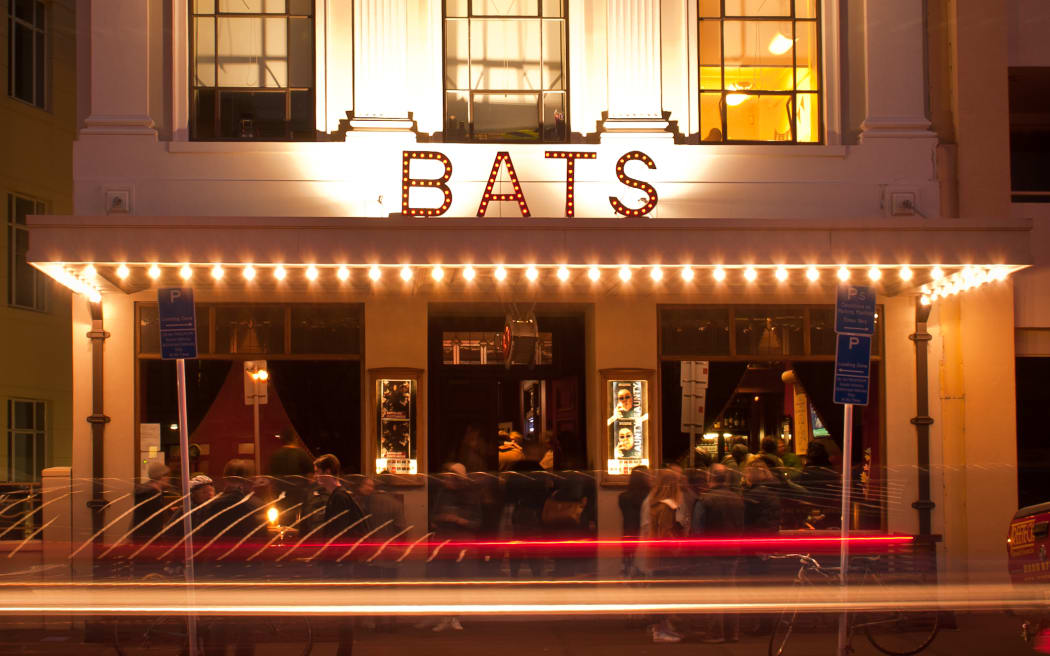 The BATS theater building from the street side at night.  The facade of the old building shines in golden light.  The word bats is written in lights.  People are inside and having a good time.