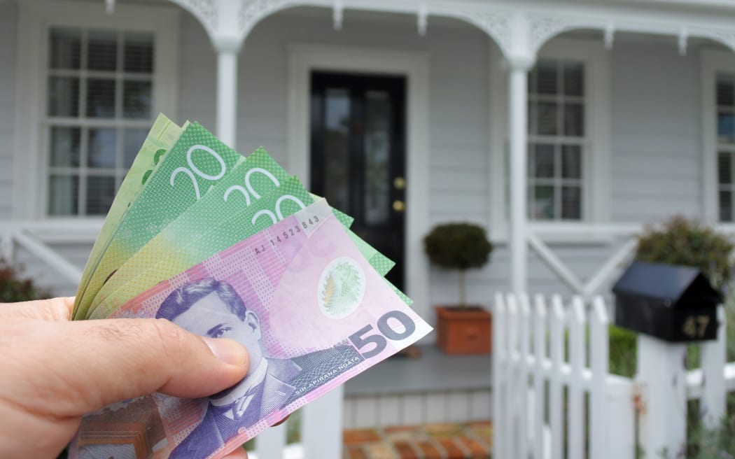 A mans hand holds NZ dollar bills against a front of a traditional villa house in Auckland, New Zealand. Buy, sale, real estate, insurance, mortgage, bank loans and housing market concept.
