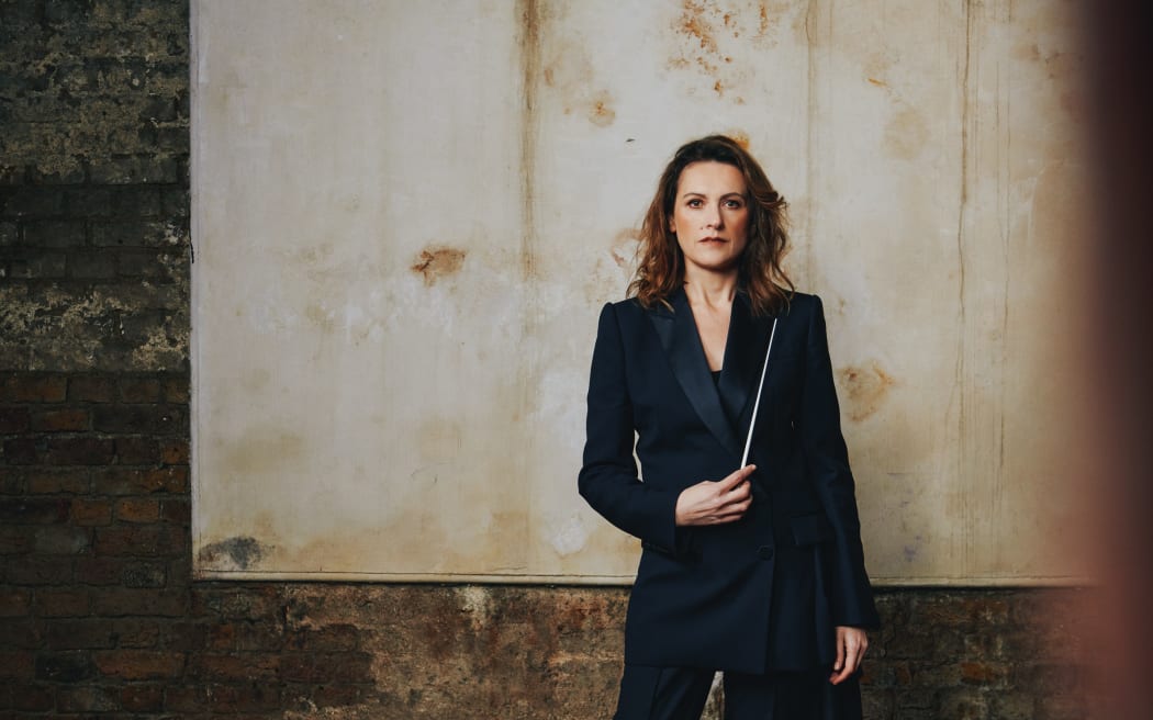 Australian conductor Natalie Murray Beale wears a black suit and stands holding the white conducting baton pointing upwards from the waist.