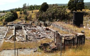 The remains of a demolished red brick building at Prohibition Mine in Waiuta, surrounded by grass and shrubs. The former mine on the West Coast is rated as the most toxic site in New Zealand