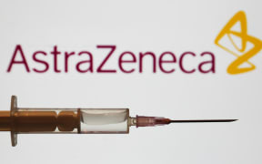 Medical syringe is seen with AstraZeneca company logo displayed on a screen in the background.