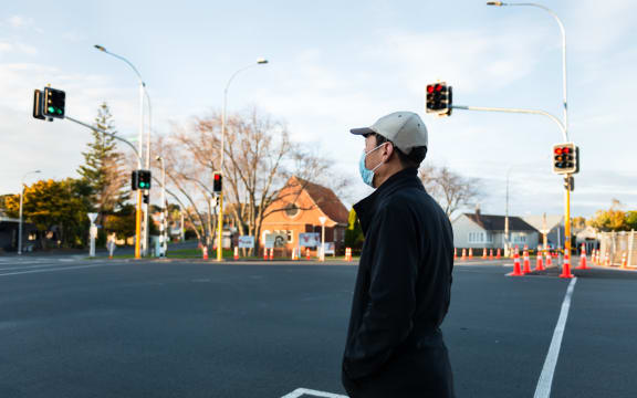 Man wearing a facemask waiting at the intersection for the green pedestrian light. Roadwork traffic cones along the street.